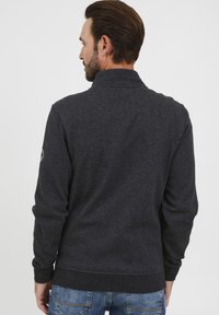 Mørk grå genser med høy krage, strukturert stoff og ribbestrikkede mansjetter. Har et logo-patch på venstre erme. Avslappet passform.