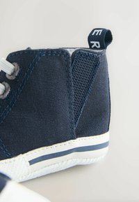 Temno modni platneni čevlji z visokim zgornjim delom in teksturiranimi stranskimi paneli, belim podplatom in kontrastnim modrim trakom. Imajo sive vezalke in zanko za poteg.