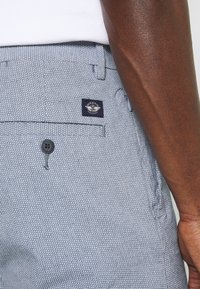 Szare spodnie w wzory z teksturowanej tkaniny, z zapięciem na guzik i granatową metką marki na tylnej kieszeni.