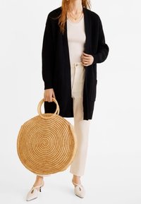 Cardigan noir en maille, top beige, pantalon blanc et chaussures beiges à bout pointu, tenant un grand sac rond en paille tressée avec un motif en spirale.