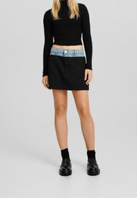 Mini jupe noire avec une taille en denim, dotée d'une fermeture à boutons à l'avant. Associée à un haut noir ajusté à manches longues et des chaussures noires.