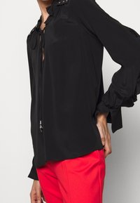 Blouse noire au design fluide, dotée d'un décolleté avec cravates et de clous décoratifs, manches longues avec des poignets à volants. Portée avec un pantalon rouge.