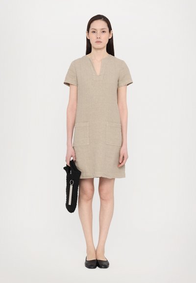 Beige linnen jurk met korte mouwen, V-hals en twee voorzakken; model houdt een zwarte stoffen tas vast met een zilveren ringdetail.