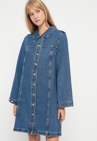 Denima kleita ar klasisku apkakli, garām piedurknēm un pogājamu priekšu ar sudraba aparatūru, vidēji zilā izskata un brīvā piegriezumā.