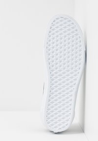 Sole bílé tenisky s šestiúhelníkovým vzorem, umístěná svisle proti bílé rohové zdi.