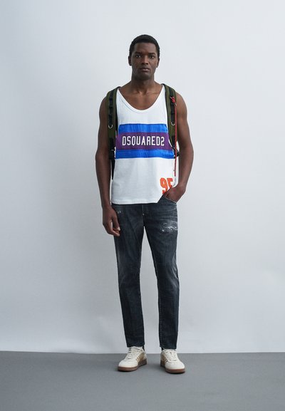 Jeune homme debout, portant un débardeur blanc avec le logo "DSQUARED2", un jean sombre déchiré, des baskets blanches et un sac à dos sur fond uni.