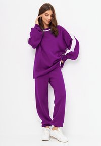 Sweatshirt violet avec des manches surdimensionnées, arborant des rayures blanches. Associé à un pantalon de survêtement violet ajusté. Des baskets blanches complètent la tenue.
