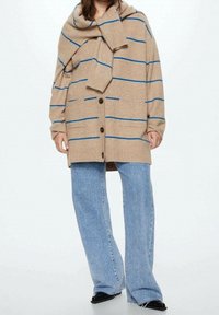 Beżowy sweter na drutach z niebieskimi paskami, z kołnierzem typu szalowego, guzikami z przodu i kieszeniami z przodu. Połączony z jasnoniebieskimi dżinsami.