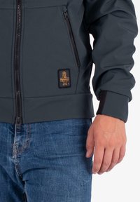 Giacca zip-up grigio scuro con un design resistente, caratterizzata da una piccola patch con logo e polsini elastici. Indossata sopra jeans di denim blu.
