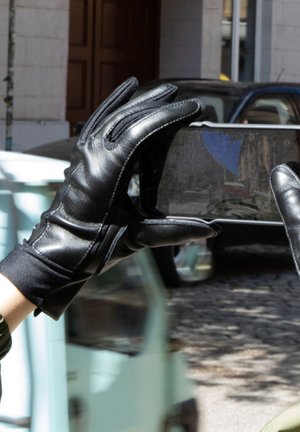 Gants en cuir noir au toucher lisse, tenus pendant la prise d'une photo sur un smartphone. L'arrière-plan montre un cadre urbain flou et des véhicules.