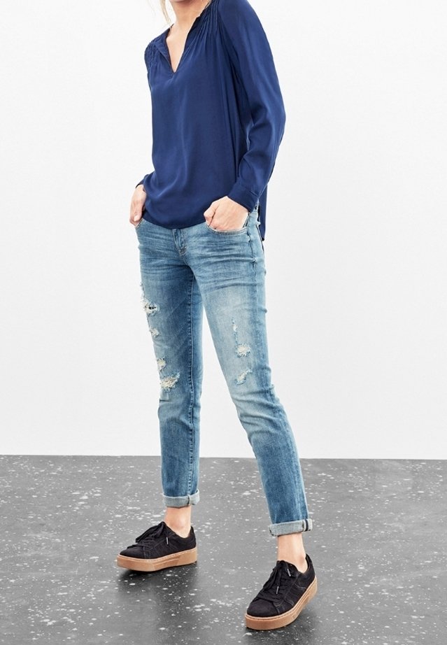 Mörkblå blus med lång ärm och veckade detaljer, matchad med urtvättade ljusblå jeans och svarta sneakers med ljusbrun sula.