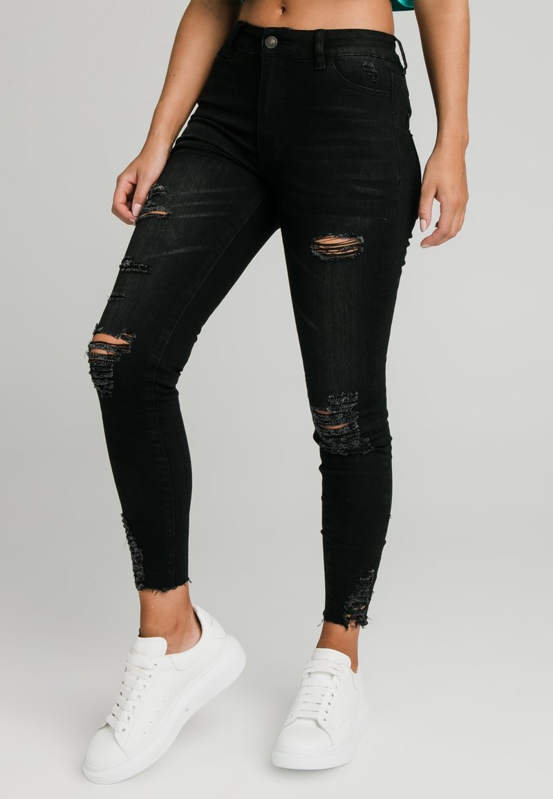 Svarta högmidjade skinny jeans med slitdetaljer. Har fransade fållar och rivna accenter. Parat med vita sneakers.