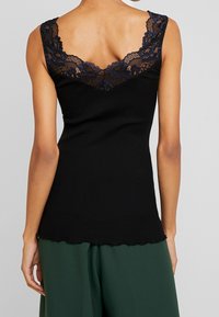 Czarny prążkowany top na ramiączkach z falowanym dołem i koronkowymi zdobieniami wzdłuż szerokiego dekoltu i ramion, noszony z zielonymi spodniami.
