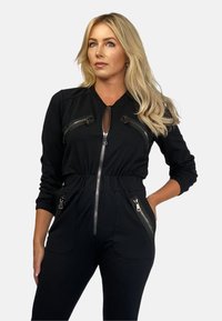 Czarny kombinezon z długimi rękawami, elastycznym pasem, kieszeniami na zamek i zamkiem błyskawicznym z przodu. Gładki materiał z dopasowanym krojem.