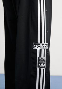 Svarta träningsbyxor med vita tre ränder längs sidan. Har Adidas-logotyp och texten "MÄRKEN MED TRE RANDER" på märket.