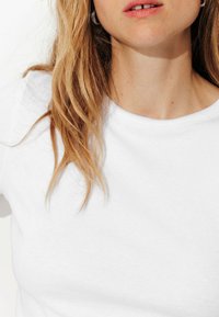 T-shirt en coton blanc avec un col rond. La mannequin a des cheveux blonds légèrement ondulés, aux épaules. Des boucles d'oreilles en argent visibles et un maquillage minimal.