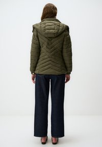 Olijfgroen pufferjack met een chevronpatroon, capuchon en geribbelde textuur, gecombineerd met wijde donkerblauwe jeans en zwarte schoenen.