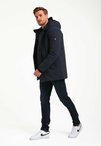 Chaqueta con capucha navy hecha de tela suave; cuenta con un cierre frontal, bolsillos laterales y un acento de logotipo en la manga. Se lleva con jeans oscuros y zapatillas.