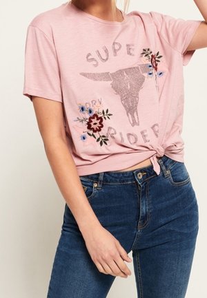 Kvinde iført en lyserød T-shirt med knude, hvor der står "Super Rider", med en grafisk tyrskalle og blomsterbroderi, kombineret med blå jeans.