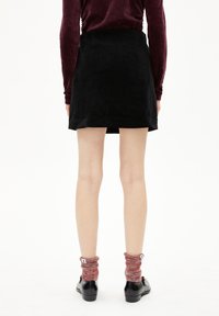 Schwarzer Wildleder-Minikilt mit geradem Schnitt, kombiniert mit roten gemusterten Socken und schwarzen Schuhen; einfarbiger Hintergrund.