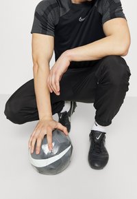 Camiseta atlética negra con acentos grises texturizados, pantalones negros, zapatillas negras y un balón de fútbol gris con franjas blancas sostenido en el suelo.