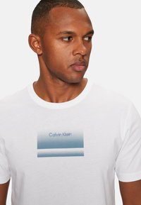 Camiseta de algodón blanca con un gráfico rectangular en degradado azul y el texto "Calvin Klein" en una fuente moderna, cuello redondo y mangas cortas.