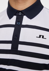 Pikétröja med marinblå och vita horisontella ränder, en ribbad marinblå krage, knapplist med tre knappar och en liten svart logotyp på bröstet.