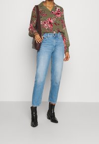 Blusa verde floreale, tessuto leggero, maniche lunghe; jeans azzurri a vita alta, gamba dritta; stivali neri alla caviglia; piccola borsa marrone.