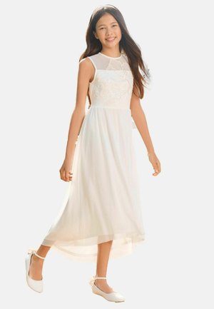 Jeune fille souriante, portant une robe blanche sans manches avec broderie florale et décolleté transparent, associée à des ballerines blanches à bride cheville.