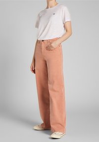 Camiseta oversized de color rosa claro con un pequeño logo negro, combinada con jeans de mezclilla color durazno claro de corte suelto, que cuentan con cintura alta y pierna recta.