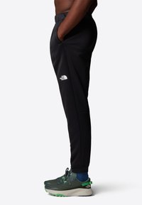 Black athletic pants with a slim fit, featuring side pockets and a logo on the lower leg. Worn with dark green sneakers and blue socks.