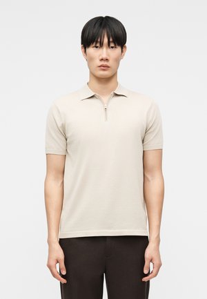 Tânăr cu păr negru, purtând un tricou polo bej cu mâneci scurte și fermoar și pantaloni închiși la culoare, stând pe un fundal simplu, deschis la culoare.