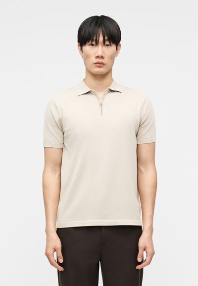 Jeune homme aux cheveux noirs portant un polo beige à manches courtes avec une fermeture éclair et un pantalon foncé, debout devant un fond clair uni.