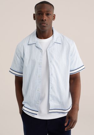 Fiatal férfi világoskék mintás rövid ujjú ingben, sötét csíkokkal az ujjakon és az alsó szegélyen, fehér póló fölött, egyszerű háttér előtt állva.