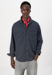 Temno modra denim srajca z dolgimi rokavi, dvema sprednjima žepoma, zapenjanjem na gumbe in okroglim robom. Nosijo se čez rdečo bandano.