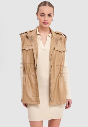 Femme portant une robe en maille côtelée beige et un gilet sans manches en cuir marron clair avec des poches avant, se tenant devant un fond uni.