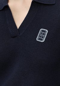 Pull en maille bleu marine avec col en V et un logo rectangulaire blanc représentant un crocodile sur la poitrine gauche.