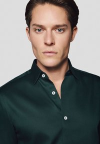 Ciemnozielona koszula z guzikami, wykonana z gładkiego materiału, z klasycznym kołnierzykiem i subtelną fakturą. Guziki są białe z delikatnym połyskiem.