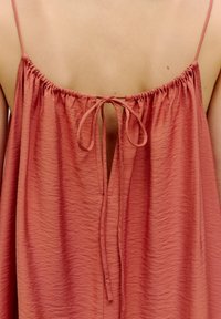 Rückansicht eines ärmellosen, rostfarbenen Kleides aus strukturierter Stoff. Verfügt über einen verstellbaren Bindeverschluss am Halsausschnitt und geraffte Nähte.