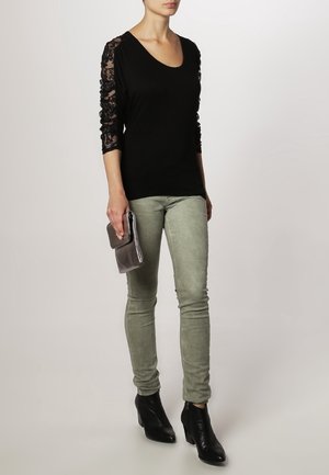 Haut noir avec des manches en dentelle, associé à un pantalon ajusté vert clair. Le modèle tient une pochette argentée et porte des bottines noires.