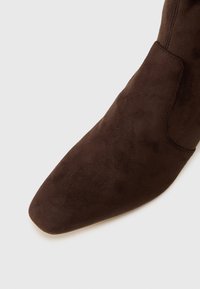 Bota de ante marrón con puntera cuadrada y costuras visibles sobre un fondo blanco.