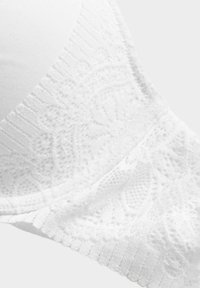 Bra bianco con tessuto lisci e dettagli in pizzo elaborati, accompagnato da un elastico testurizzato. Il pizzo presenta un motivo floreale, arricchendo l'interesse visivo.
