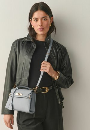 Fiatal, sötét hajú nő fekete bőrdzsekit, fekete felsőt, arany fülbevalót és karkötőt visel, világosszürke táskát tart arany színű díszítéssel.