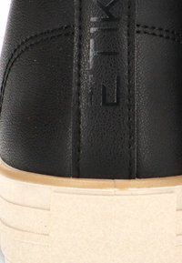 Acercamiento del talón de una zapatilla deportiva de cuero negro texturizado con una tira de logo vertical y una gruesa suela de goma beige.
