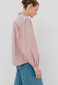 Donna con un chignon che indossa una blusa lunga e ampia rosa e jeans blu, in piedi contro uno sfondo chiaro uniforme, mentre guarda verso il basso.