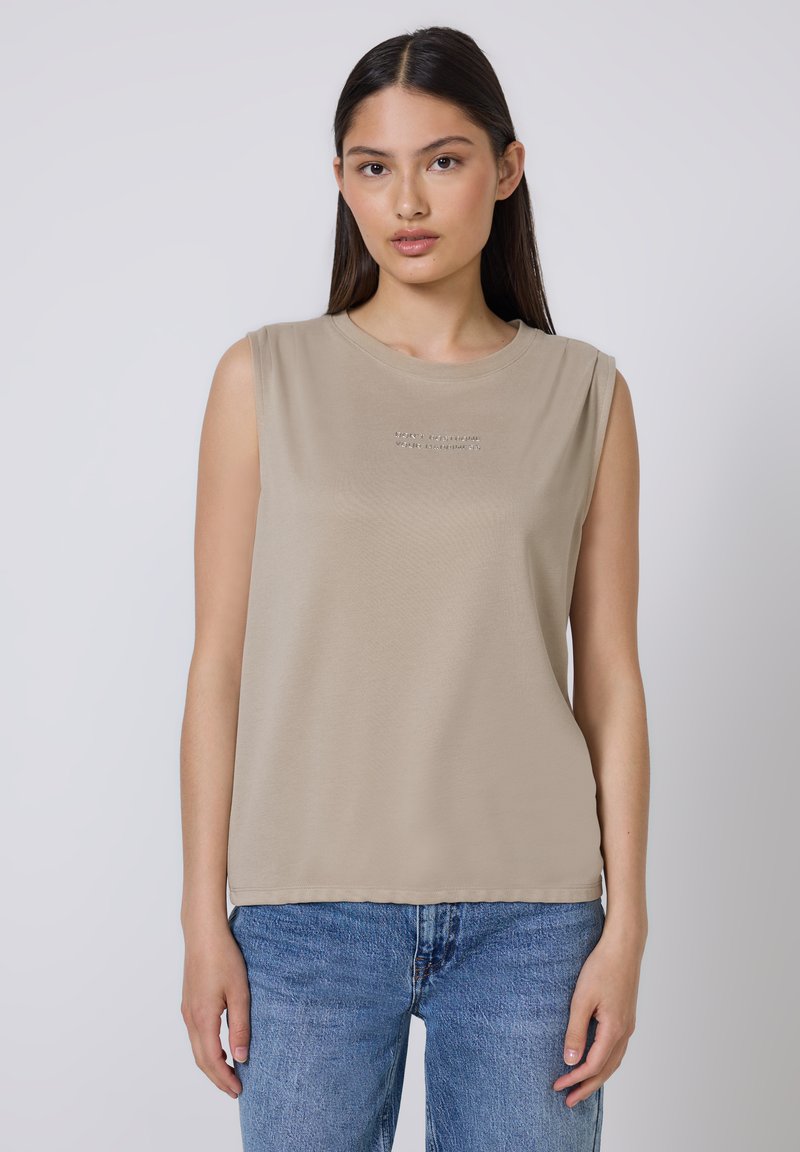 Top sin mangas beige hecho de una tela suave, con un escote redondo y un sutil detalle de texto en el frente. Combinado con jeans azules.