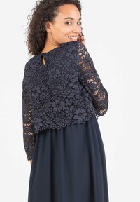 Marineblaues Kleid mit einem Spitzentop mit floralen Mustern und langen Ärmeln sowie einem Schlüsselloch-Detail im Rücken, ergänzt durch einen fließenden Rock.