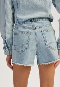 Shorts de mezclilla azul claro con dobladillo deshilachado, dos bolsillos traseros y detalles de costura visibles. Combinados con una chaqueta de mezclilla a juego.