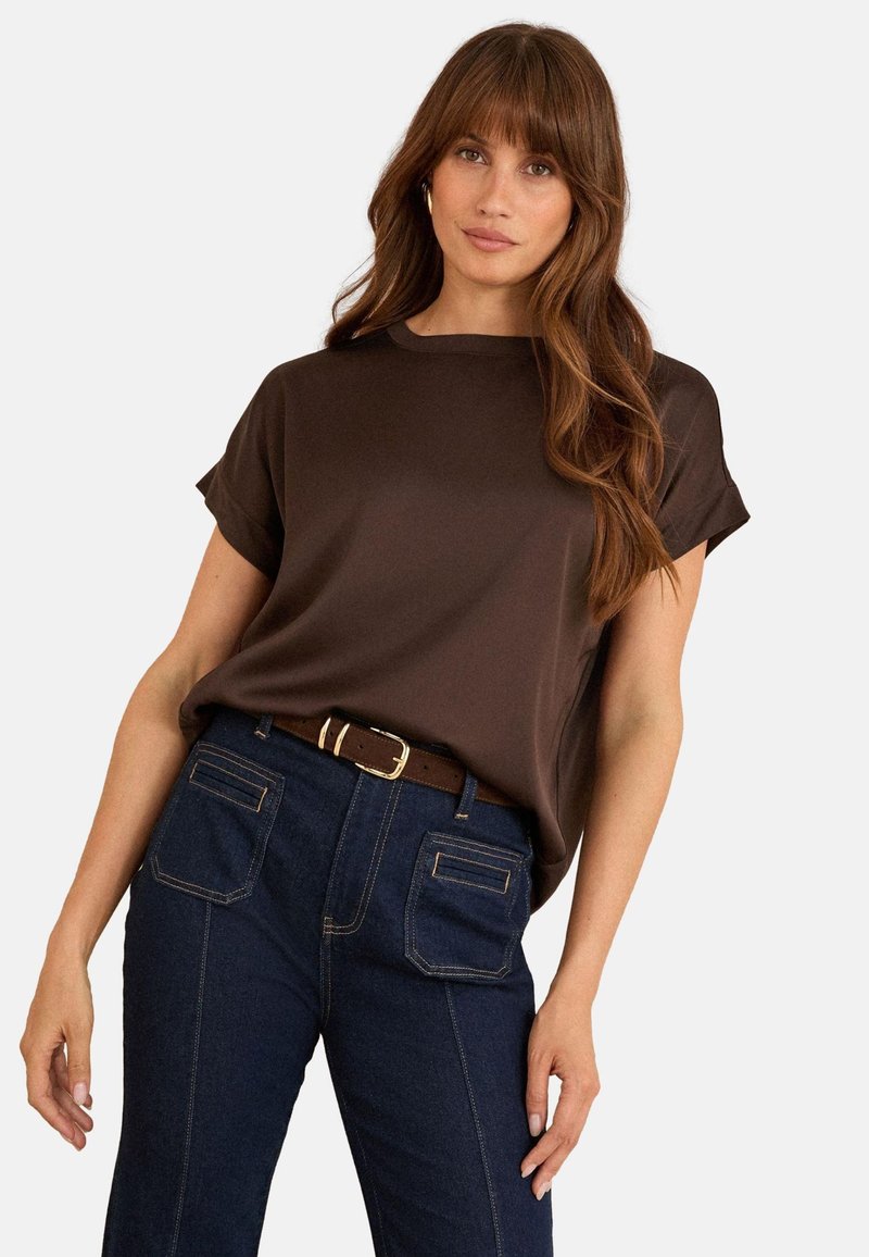 Hnědý krátký rukáv s uvolněným střihem a hladkou texturou, spárovaný s tmavě modrými džínami s vysokým pasem, zdůrazněný hnědým opaskem.