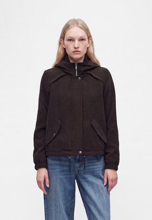 Jeune femme portant une veste zippée marron foncé avec des poches à rabat et des poignets élastiques, associée à un jean bleu ample sur un fond uni.